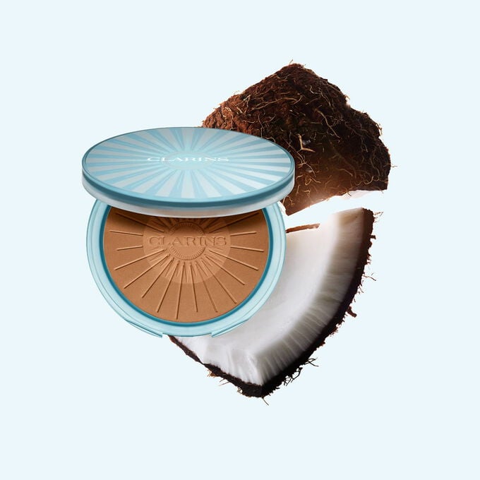 Imagem do produto Summer Bronzing Powder rodeado por cocos, ilustrando o seu ingrediente principal para um brilho natural e bronzeado.