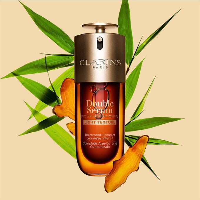 Um frasco de 70 ml de Clarins Double Serum Light Texture entre folhas de curcuma e cana-do-reino sobre um fundo laranja.