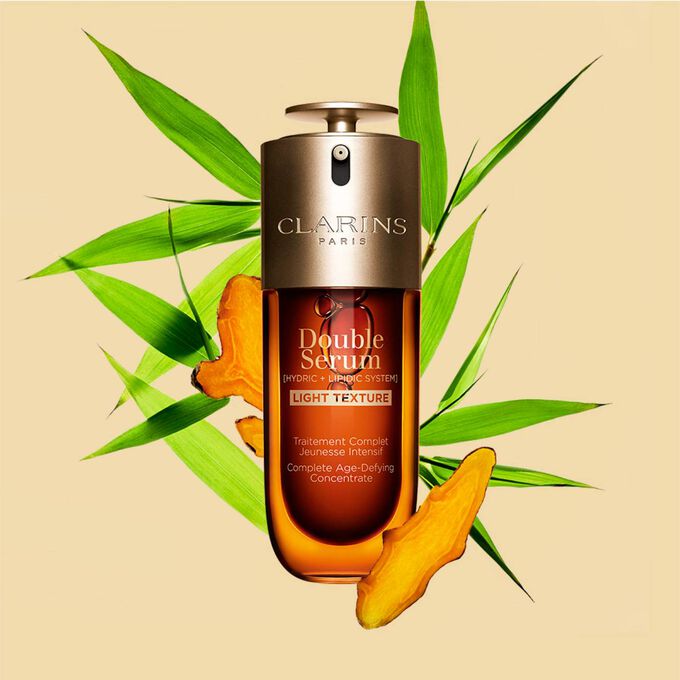 Um frasco de 50 ml de Clarins Double Serum Light Texture entre folhas de curcuma e cana-do-reino sobre um fundo laranja.