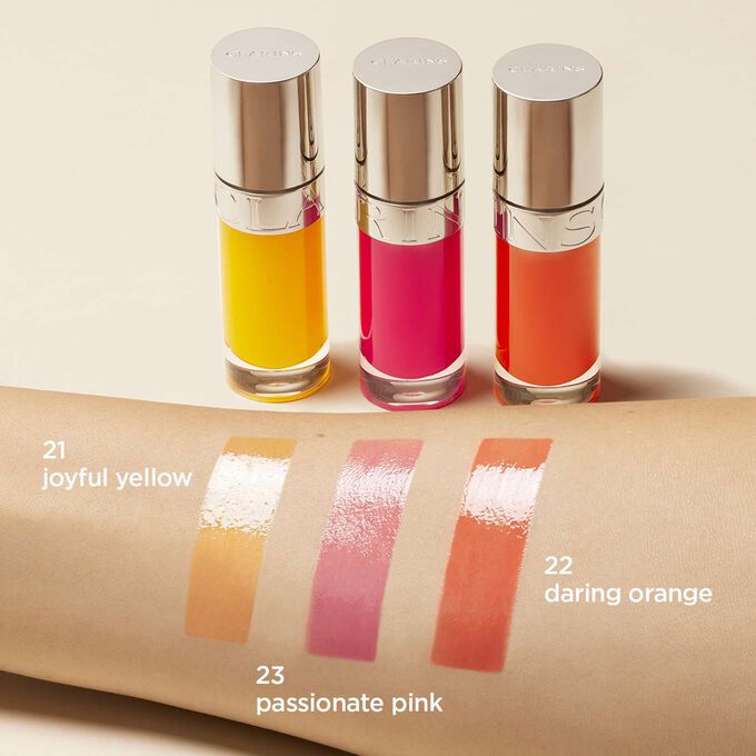 Apresentação da gama de óleos para lábios Clarins em amarelo, amarelo e laranja, com as cores aplicadas no braço de uma mulher